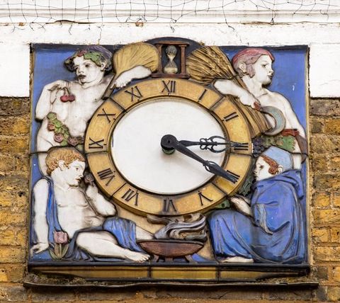 Architectural detail. A round clock face on the exterior of a building is set into a larger square, the corners of which are filled with personifications of the seasons. Children with white-coloured skin hold berries or sheaves of wheat, or sit by a fire or next to a flower. Most of the glazes are vivid blue or golden yellow, with a few touches of red and green.
